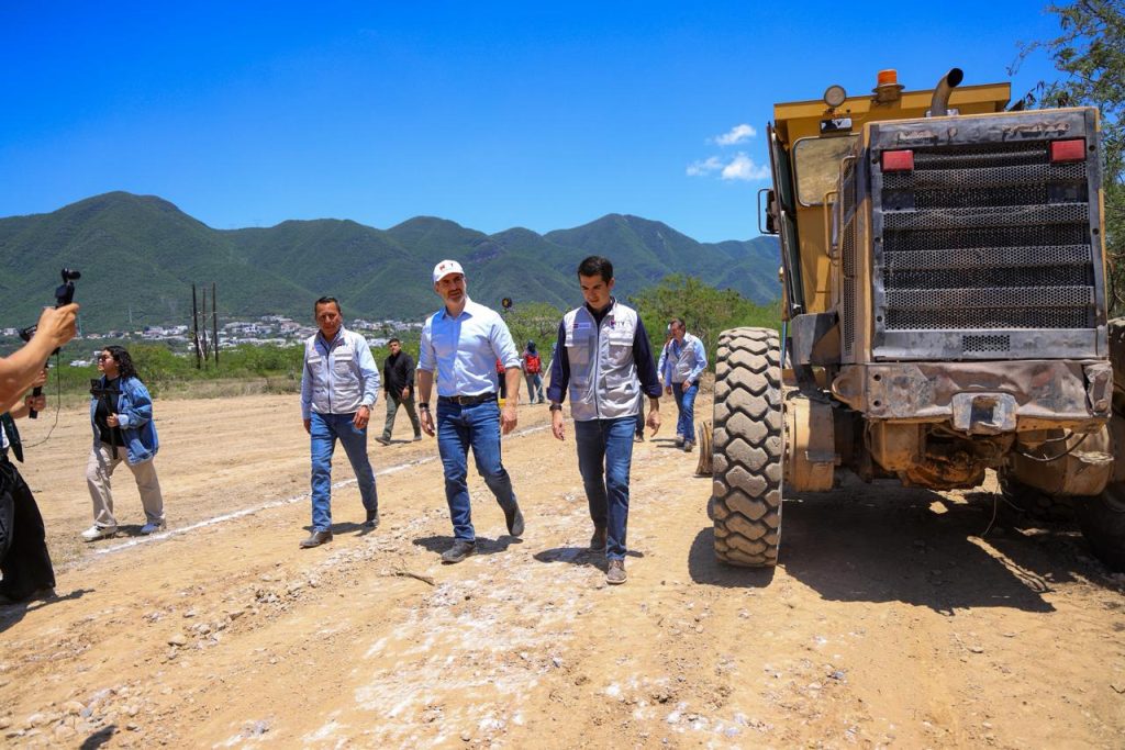 Group of Workers and Officials Walking Across a Dusty Construction Site Beside a Large Yellow Dump Truck, with Green Mountains in the Background. - Capitanes Regios