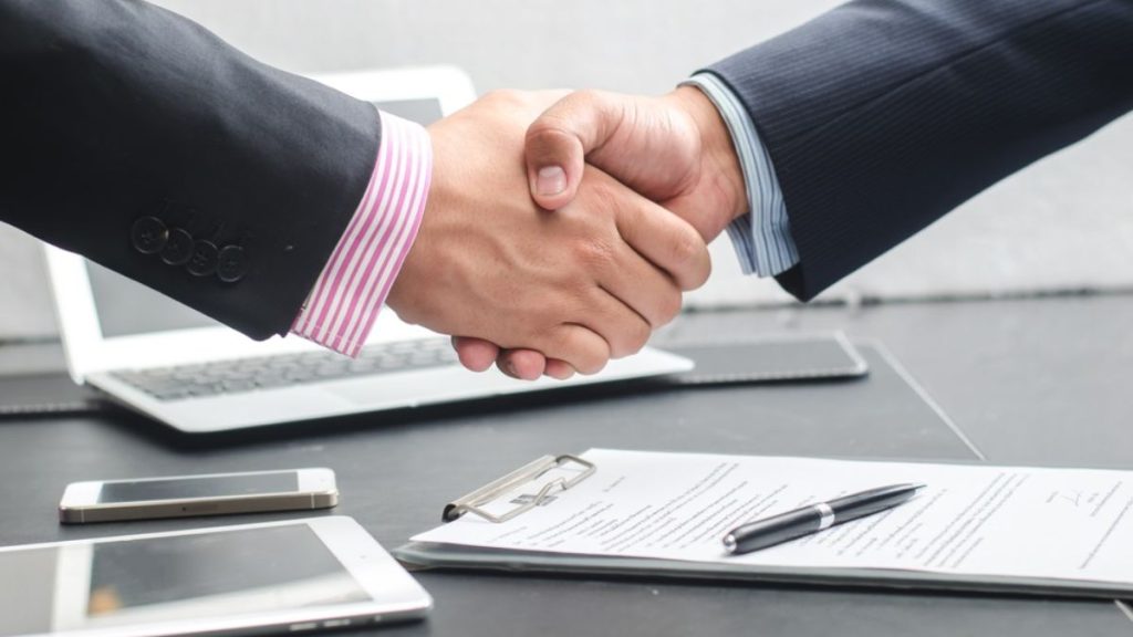 Two Professionals Shake Hands over a Desk with a Clipboard, Pen, Laptop, and Mobile Devices Nearby, Signaling a Deal. - Capitanes Regios