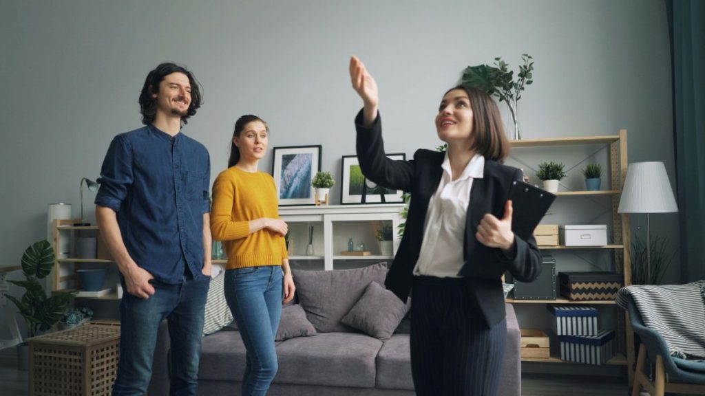 Professional Woman in a Black Blazer Raises Her Hand to Present to Two Clients in a Modern Living Room. - Capitanes Regios