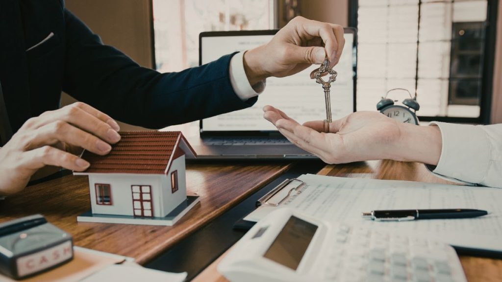 Person Hands a Key to Another Across a Desk, with a Miniature House and Documents, Signaling a Real Estate Closing. - Capitanes Regios