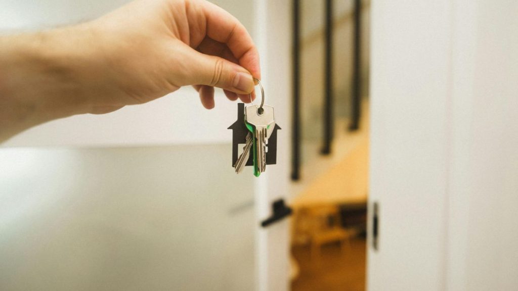 Cómo-saber-cuánto-cuesta-mi-casa-quantium-desarrollos - Capitanes Regios Hand Holds a Keyring with House-shaped Key Fobs and Several Keys in Front of an Interior Doorway, Suggesting Moving in or Home Ownership Scenarios. - Capitanes Regios
