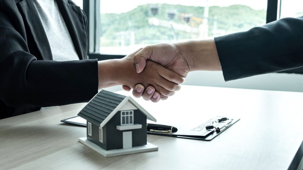 Two Professionals Shake Hands over a Desk, with a House Model and Clipboard, Signaling a Real Estate Agreement As a Deal is Made. - Capitanes Regios