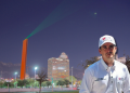 Man in a White Shirt and Cap Stands in a Nighttime Urban Square with a Tall Orange Sculpture and Green Laser Beam in the Sky Behind Him. - Capitanes Regios