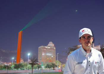 Renuevan-faro-del-comercio - Capitanes Regios Man in a White Shirt and Cap Stands in a Nighttime Urban Square with a Tall Orange Sculpture and Green Laser Beam in the Sky Behind Him. - Capitanes Regios