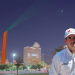 Renuevan-faro-del-comercio - Capitanes Regios Man in a White Shirt and Cap Stands in a Nighttime Urban Square with a Tall Orange Sculpture and Green Laser Beam in the Sky Behind Him. - Capitanes Regios