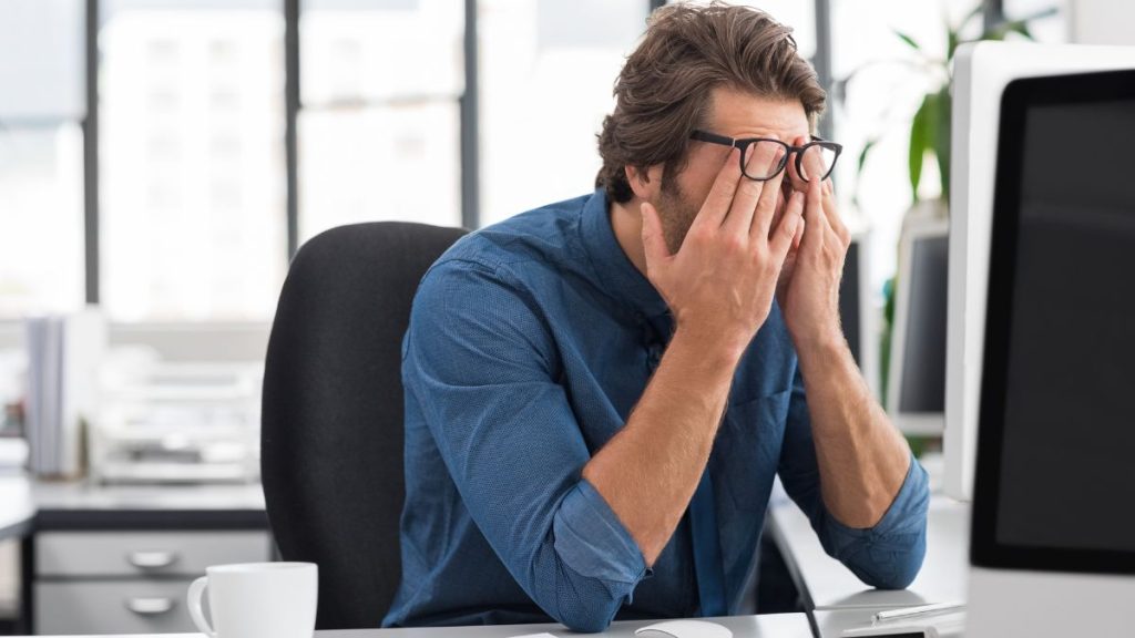 Man in a Blue Shirt Sits at a Desk in an Office, Rubbing His Eyes in Frustration While Looking at a Computer Screen. - Capitanes Regios