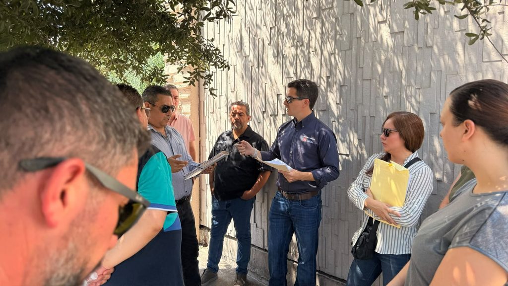 Group of Coworkers Standing Outdoors, Reviewing Papers and Discussing a Document Under a Tree-shaded Area Near a Textured Wall. - Capitanes Regios