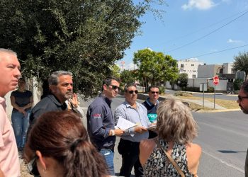 Group of People Standing on a Sunny Street Corner, Holding Papers and Talking, with Trees and a Stop Sign in the Background. - Capitanes Regios