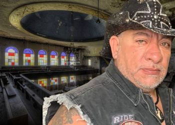 Man wearing a studded black cowboy hat and a sleeveless denim vest stands in a dilapidated church with colorful stained-glass windows in the background.
