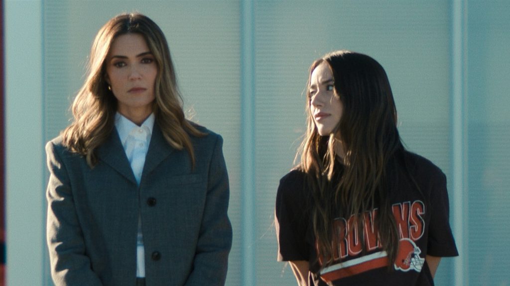 Two Women Stand Outdoors Beside a Light-colored Wall; Left Woman Wears a Gray Blazer and White Shirt, Right Woman Wears a Black Bruins Sweatshirt with Long Hair. - Capitanes Regios