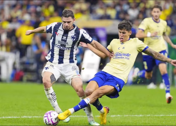 Two Soccer Players in a Blue‑and‑white Striped Kit and a Yellow Kit Fight for the Ball on a Green Field During a Match. - Capitanes Regios