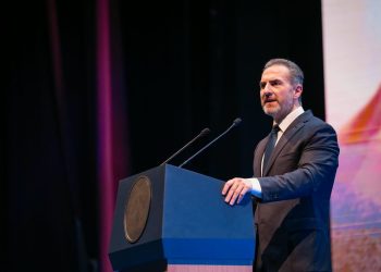 Man in a Dark Suit Delivering a Speech at a Blue Podium on Stage with Two Microphones Behind Him. - Capitanes Regios