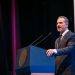 Man in a Dark Suit Delivering a Speech at a Blue Podium on Stage with Two Microphones Behind Him. - Capitanes Regios