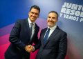Two Men in Dark Suits Shake Hands and Smile at a Formal Event, with a Blue-pink Backdrop and Bold White Text Behind Them. - Capitanes Regios