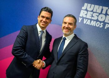 Two Men in Dark Suits Shake Hands and Smile at a Formal Event, with a Blue-pink Backdrop and Bold White Text Behind Them. - Capitanes Regios