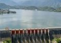 Dam Spanning a Calm Lake with Spillway Gates Releasing Water, Village on the Left, and Green Mountains in the Background. - Capitanes Regios