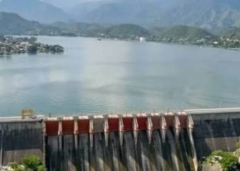 Dam Spanning a Calm Lake with Spillway Gates Releasing Water, Village on the Left, and Green Mountains in the Background. - Capitanes Regios