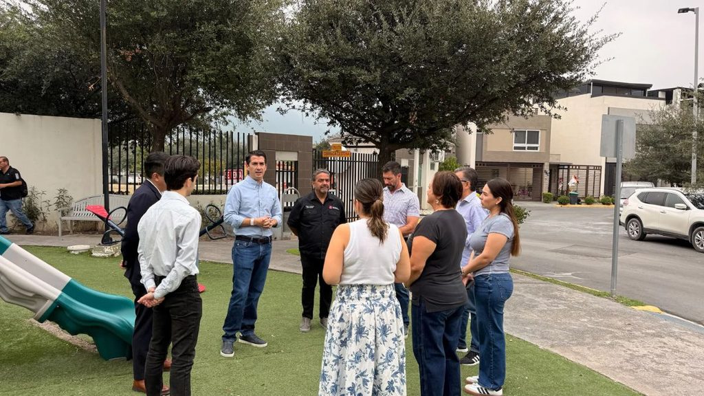Group of Adults Standing in a Circle Outdoors, Having a Discussion Near a Playground and Modern Buildings Behind Them. - Capitanes Regios